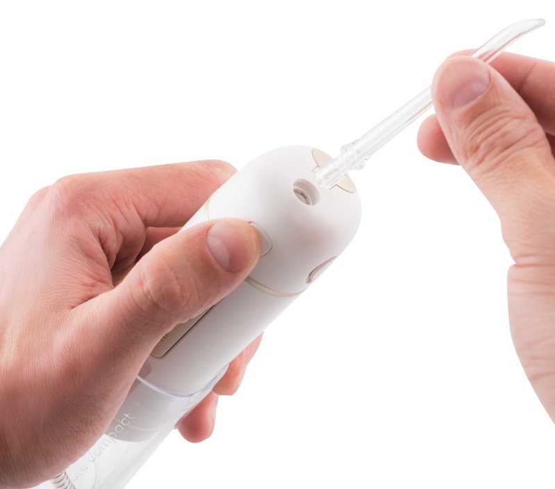 Detailní záběr rukou, které drží bílou ústní sprchu ETA Aqua Care Compact a nasazují do horního otvoru průhlednou výměnnou trysku. Obrázek demonstruje snadnou manipulaci a přípravu přístroje k použití