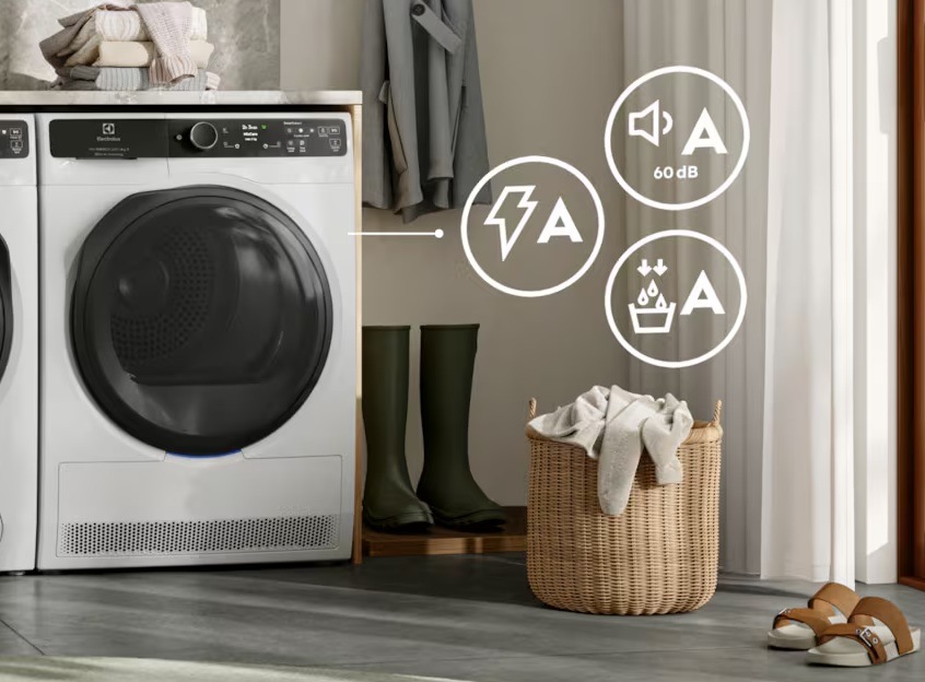 Sušička Electrolux, vpravo symboly pro energetickou třídu A. Vpravo u sušičky jsou postavené gumáky a koš na prádlo.