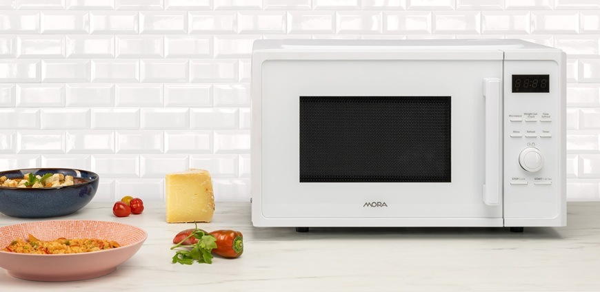 Na obrázku je Mora MT423W v bílém provedení s pootevřenými dvířky. Mikrovlnka je položená na kuchyňské lince a kolem ní jsou potraviny, které v ní lze ohřát, uvařit nebo rozmrazit.