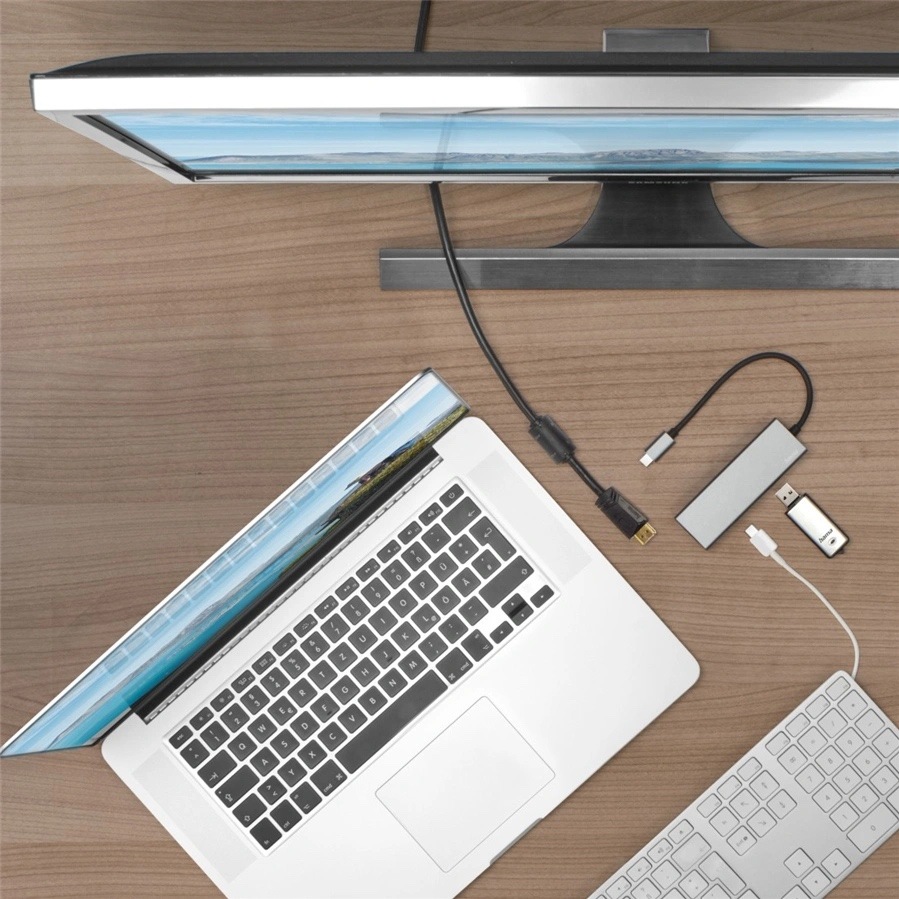 USB Hub Hama Multiport šedý Na obrázku je pracovní stůl z pohledu shora. Uprostřed leží otevřený stříbrný notebook, nahoře je velký monitor a vpravo dole bílá klávesnice. Vedle notebooku je položený šedý USB-C hub Hama s krátkým kabelem a několik dalších kabelů a malých adaptérů. Fotka ukazuje běžné použití hubu v pracovním prostředí při připojení notebooku k monitoru a dalšímu příslušenství.