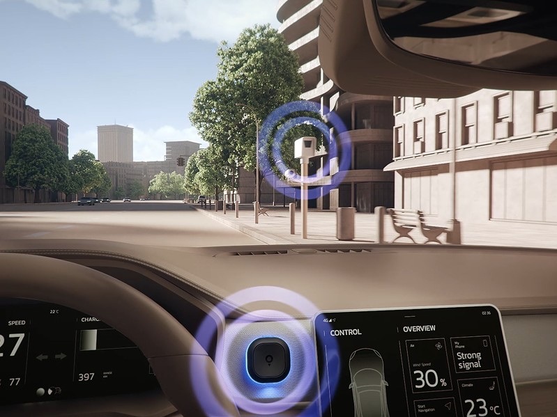 Pohled z interiéru automobilu na městskou ulici. Cestovní asistent TomTom Tom je upevněný na palubní desce vedle dotykového displeje a modře svítícím LED kroužkem upozorňuje na rychlostní kameru před vozidlem.