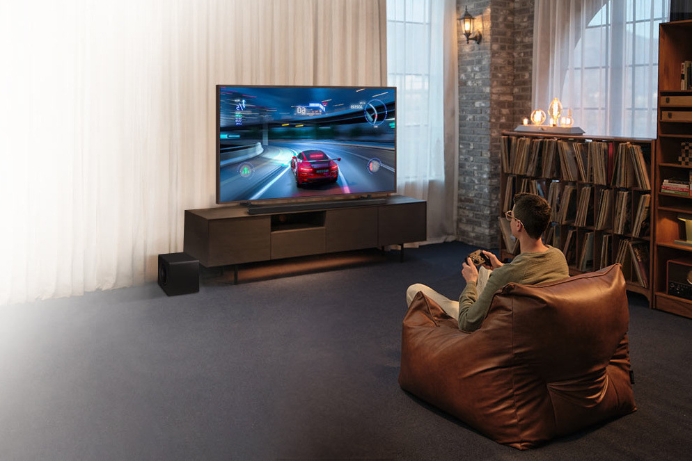 Muž sedí v sedacím vaku na zemi před televizí a hraje videohru, pod televizí leží soundbar Samsung HW-S800D