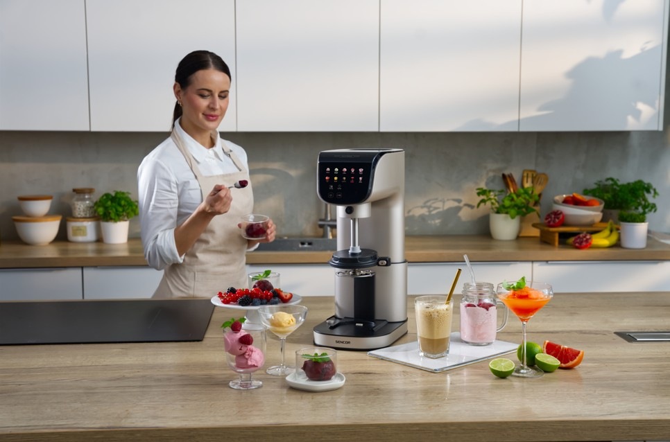 Zmrzlinovač Sencor SIC 5050WS GELATERIA champagne Žena připravuje domácí dezert pomocí zmrzlinovače Sencor SIC 5050WS GELATERIA v moderní kuchyni. Na pracovní desce jsou naservírované poháry se zmrzlinou, čerstvé ovoce a osvěžující nápoje, které dotvářejí atmosféru letního pohoštění. Elegantní přístroj s dotykovým displejem stojí uprostřed scény.