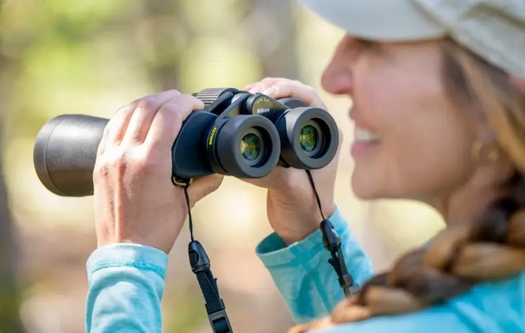 Dalekohled Nikon Action 7×50 černý Usměvavá paní drží v ruce dalekohled Nikon Action 7×50 a dívá se před sebe na krajinu.