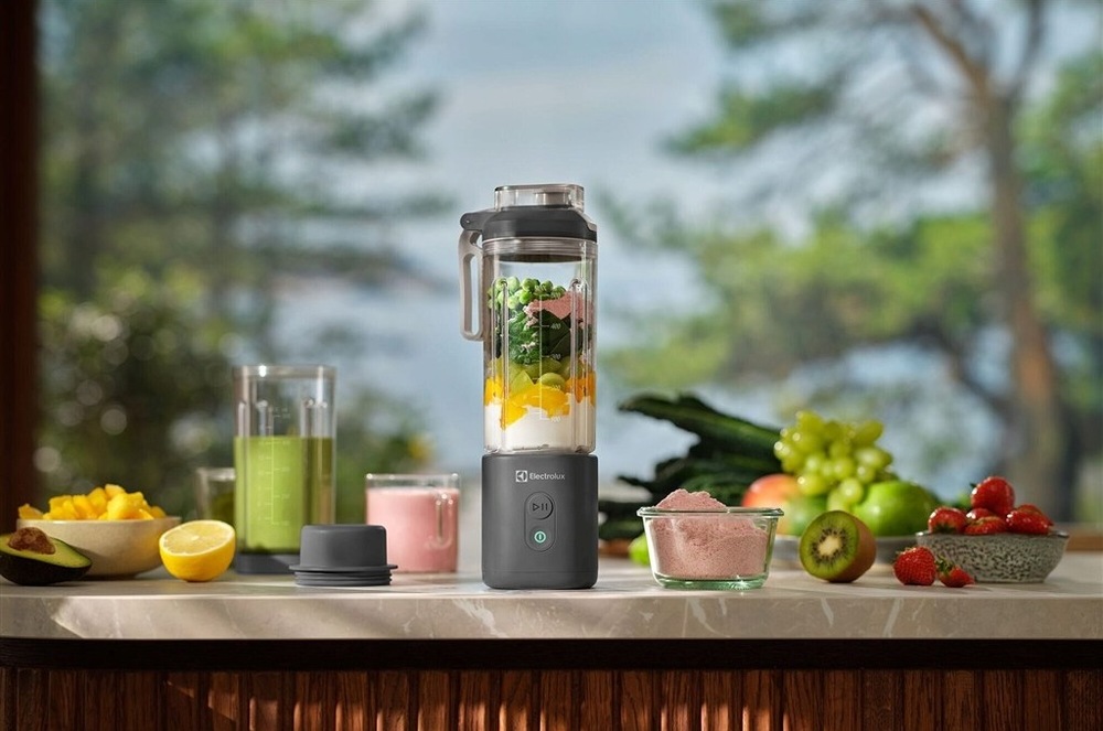 Stolní AKU mixér Electrolux E4PB1-2CG na kuchyňské lince. Uvnitř mixéru je směs pro smoothie. Na lince v pozadí jsou misky s ovocem.