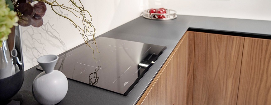 Detailní pohled na minimalistickou indukční desku ETA 679290000 v černé barvě, zasazenou do grafitové pracovní plochy s dřevěnými skříňkami v pozadí