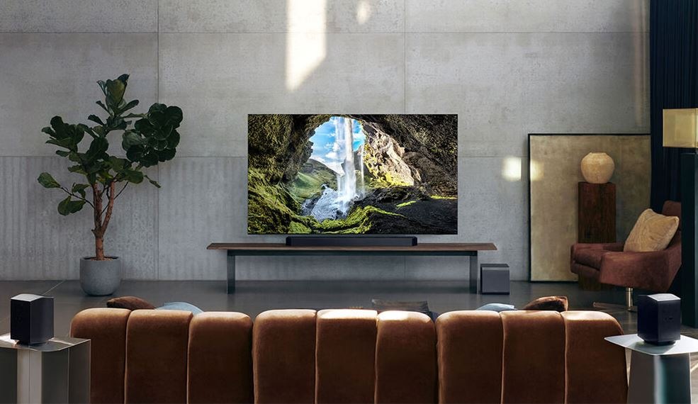 soundbar samsung pod televizi samsung v moderním obyvacím pokoji. Vedle TV je rostlina a naproti gauč. V telvizi je promítnutá příroda, skály a padající voda.