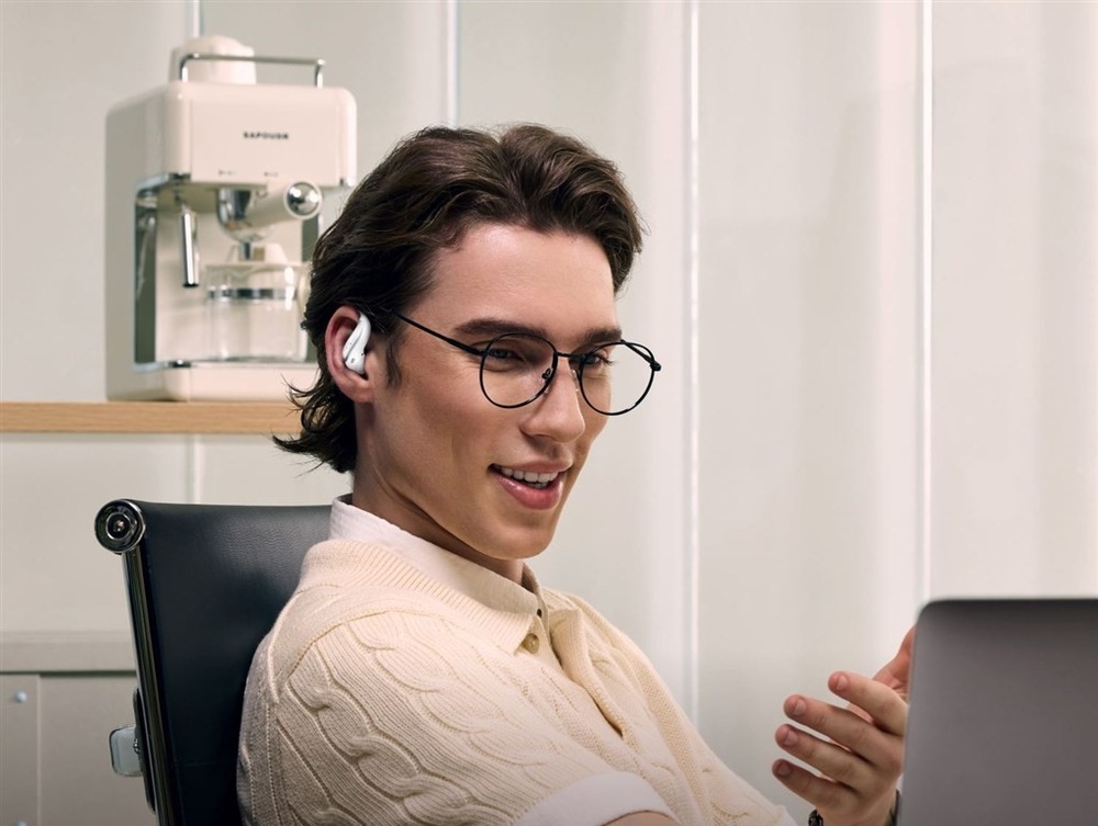 Sluchátka Shokz OpenFit Air 2, Bluetooth bílá Muž v brýlích sedí v kanceláři u pracovního stolu, má nasazené sluchátka OpenFit Air 2, v ruce drží telefon a s někým hovoří.
