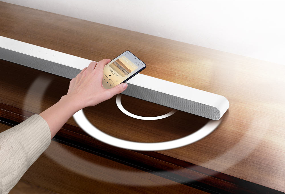 Soundbar Samsung HW-S701D bílý Ruka drží mobilní telefon přehrávající hudbu nad bílým soundbarem Samsung HW-S701D, z nějž vycházející bílá kolečka znázorňující zvuk