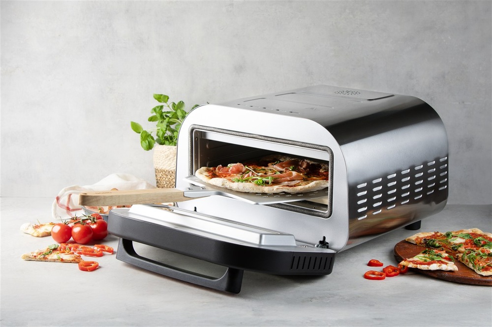 Pec na pizzu Domo GENIUS DO9289PZ je otevřená, do pece je vložena lopatka, na níž leží pizza, v okolí pece leží ingredience a hotová pizza