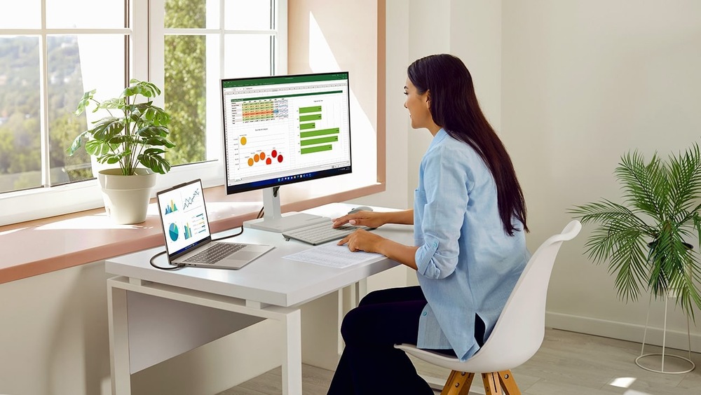 Monitor Lenovo L27h-4A šedý Žena pracuje u stolu s notebookem a monitorem, na kterém jsou zobrazeny tabulky a grafy. Světlá místnost s velkými okny, rostlinami a klidnou, soustředěnou atmosférou.