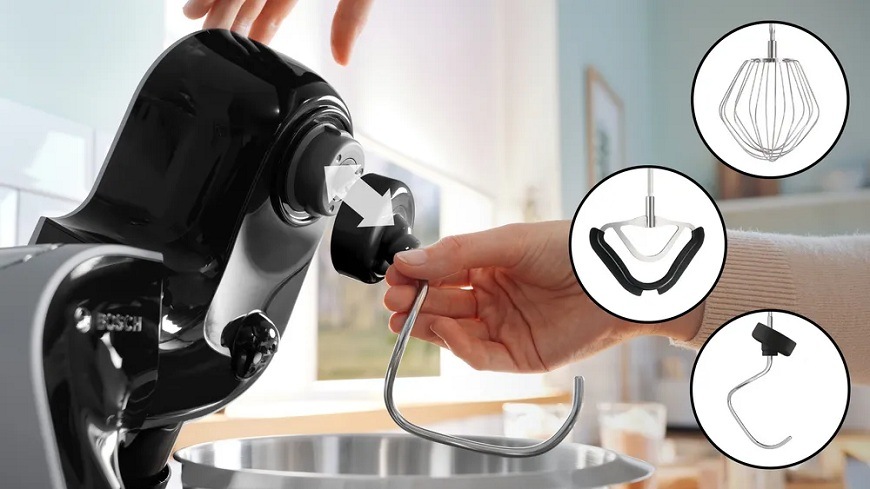 Detailní záběr ruky, která připevňuje hnětací hák do hlavy černého kuchyňského robotu.Bosch MUM5XC74. V pravé části jsou v kruzích ukázány další nástavce