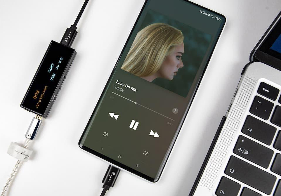 Sluchátkový zesilovač Cayin RU6 černý Na fotce je přenosný DAC převodník Cayin RU6 připojený k mobilnímu telefonu a vedle leží část notebooku. V telefonu je otevřený hudební přehrávač se skladbou Easy On Me od Adele, takže obrázek ukazuje poslech hudby v praxi. Černý RU6 je připojen k telefonu kabelem a z druhé strany do něj vedou sluchátka. Na malém displeji převodníku svítí technické údaje. Celá scéna působí čistě, moderně a ukazuje použití zařízení s mobilem i notebookem.