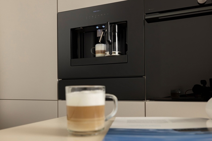 Espresso Gorenje G800 CM4G8BG černá Na stole je položen hrneček s cappuccinem. V pozadí je zabudovaný kávovar Gorenje CM4G8BG, který právě dovařil kafé s bohatou pěnou..