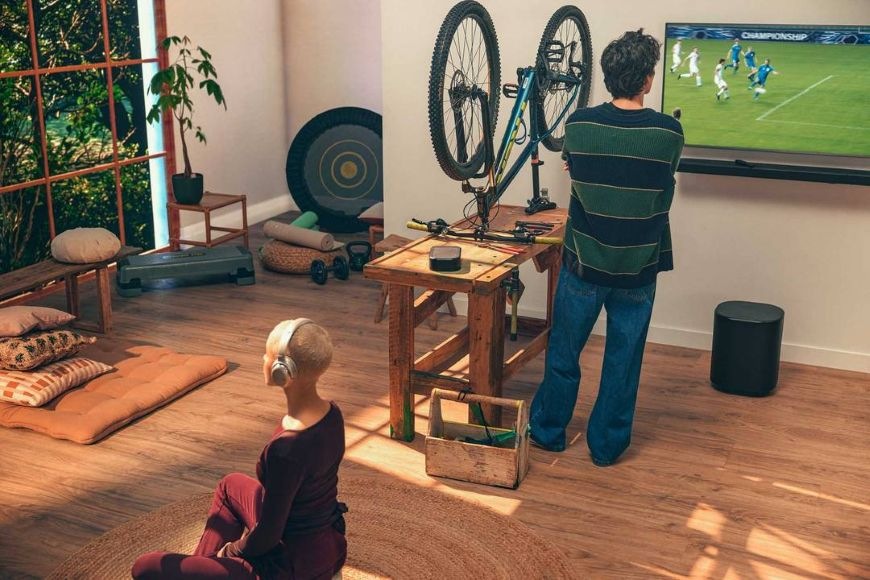 Soundbar JBL BAR 1000 MK2 černý Muž opravuje kolo a sleduje fotbal v televizi, pod kterou se nachází subwoofer JBL BAR 1000 MK2