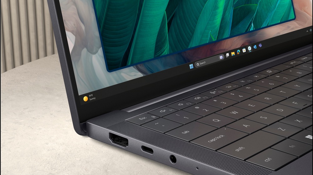 Elegantní notebook leží otevřený na světlém povrchu a na jeho zářivém displeji se zobrazují zelené listy. Stříbrné tělo je vybaveno porty USB a HDMI.