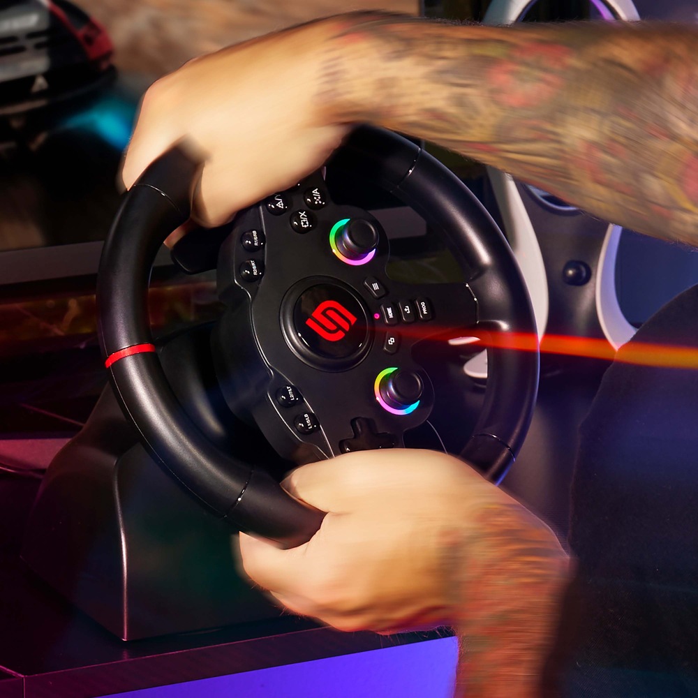 Volant SUPERDRIVE 220 RS-5 černý Tetované ruce svírají herní volant s LED podsvícením analogových páček. V pozadí je rozmazaná obrazovka závodní hry, která evokuje vzrušení a rychlost.