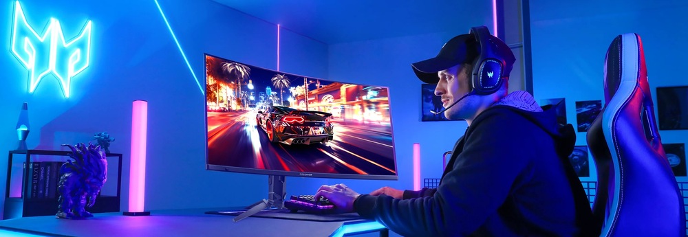 Herní monitor Acer Predator X34X5bmiiphuzx černý Hráč s nasazenými sluchátky a kšiltovkou sedí u moderního herního zařízení v modře osvětlené místnosti. Na velkém zakřiveném monitoru vidíme scénu závodní hry.