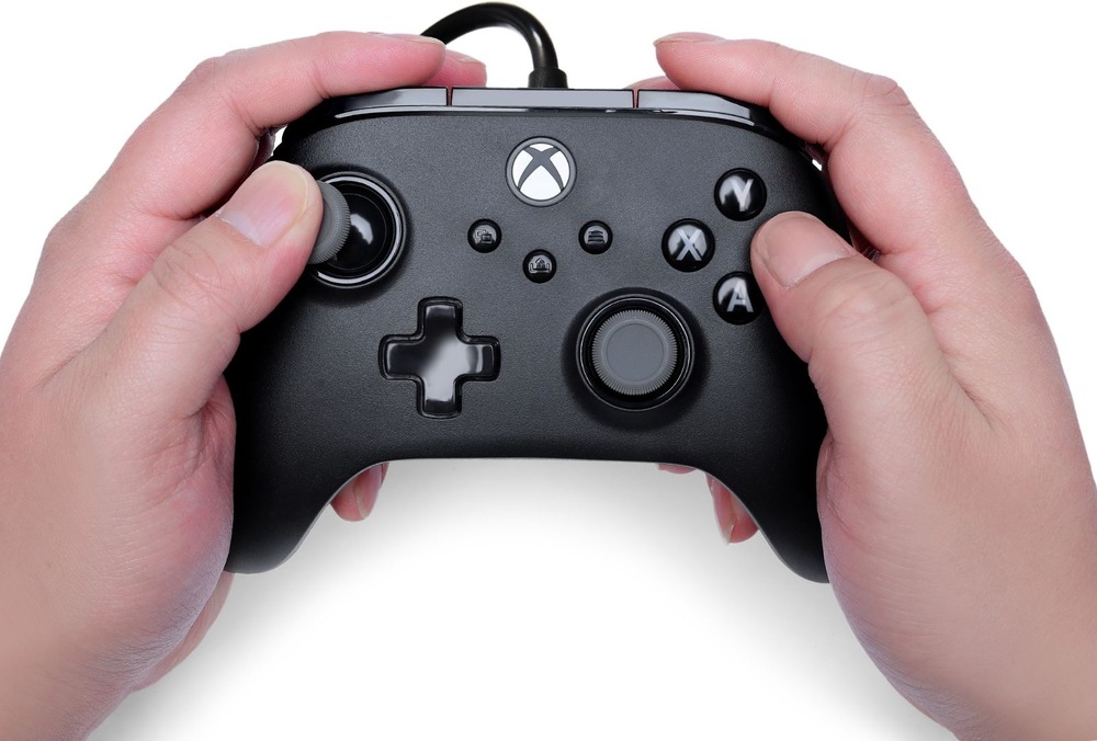 Na obrázku drží člověk oběma rukama herní ovladač PowerA Advantage Plus během hraní. Palce spočívají na analogových páčkách a tlačítkách, což ukazuje běžný způsob ovládání her. Dobře jsou vidět tlačítka A, B, X a Y, směrový kříž, funkční tlačítka uprostřed i tlačítko s logem Xbox. Snímek demonstruje ergonomický tvar ovladače, díky kterému pohodlně padne do rukou i při delším hraní.