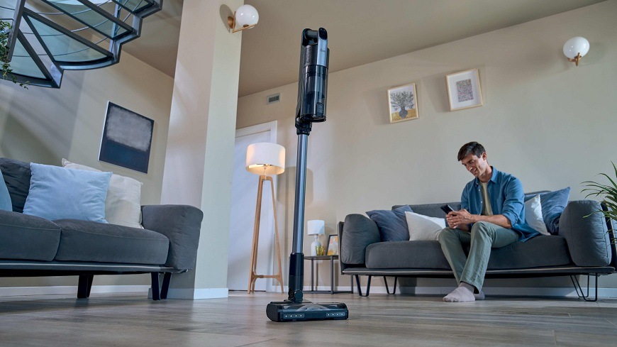 Tyčový vysavač Hoover HF310H 011 stojí uprostřed místnosti, zatímco muž na gauči používá telefon
