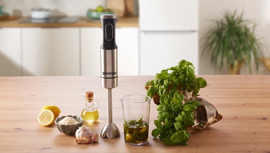 Ponorný mixér Gorenje HBX1000EB stojí na kuchyňské lince v moderní světlé kuchyni. Vedle něj je položen olivový olej, citron, bazalka a stroužky česneku.