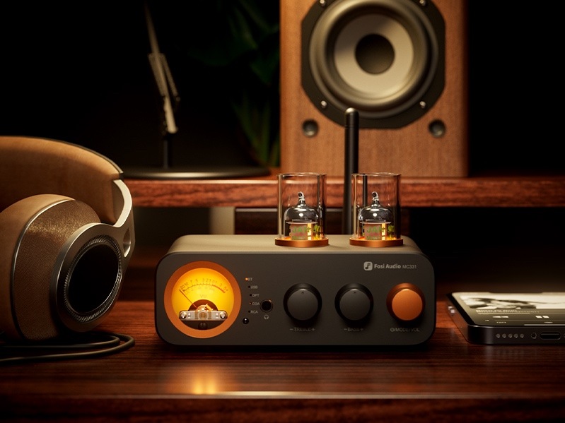 Zesilovač Fosi Audio MC331 černý Na obrázku je zesilovač Fosi Audio MC331 umístěný v domácí audio sestavě na dřevěném stole. Samotný přístroj má černé tělo, na horní straně dvě elektronky pod skleněnými kryty a na předním panelu výrazný oranžově podsvícený VU měřák. Vedle něj jsou tři otočné ovladače, z nichž pravý je oranžový. Vlevo leží velká sluchátka, v pozadí je reproduktor a vpravo mobilní telefon, takže celá scéna působí jako stylově nasvícený Hi-Fi koutek pro poslech hudby.