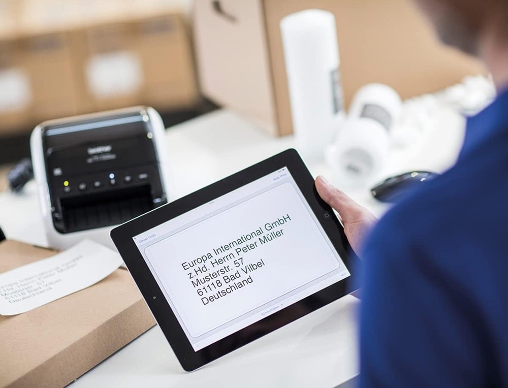 Muž drží v ruce tablet s adresou, kterou vytiskne v pozadí nacházející se tiskárna štítků Brother QL-1110NWBC.