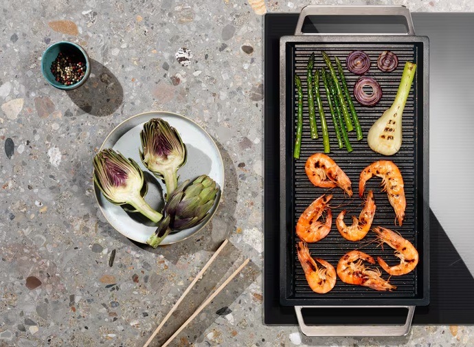 Indukční varná deska Electrolux Inspiration EIV634 černá Plancha gril s krevetami a zeleninou na spojených varných zónách. Vedle na lince talíř se zeleninou, dřevěná pinzeta a miska s pepřem.