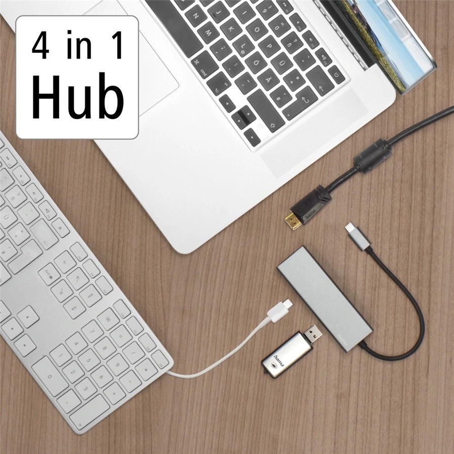 USB Hub Hama Multiport šedý Na obrázku je pracovní stůl z pohledu shora s notebookem, klávesnicí a šedým USB-C hubem Hama. Hub leží vpravo dole a je k němu připojený krátký kabel s USB-C konektorem, vedle něj jsou další kabely a malý USB adaptér. Vlevo nahoře je výrazný nápis „4 in 1 Hub“. Fotka ukazuje praktické použití hubu pro připojení příslušenství k notebooku.