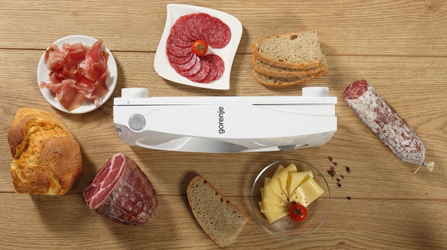 Kráječ Gorenje R401W v bílém provedení je složen na kuchyňské lince. Kolem něj jsou nakrájené nejrůznější potraviny. Nachází se tam uzeniny, chléb , maso i sýry.