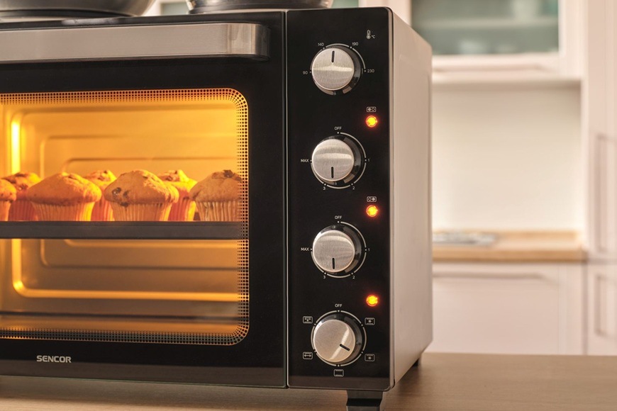Detail mini trouby Sencor SEO 4800BK s osvětleným vnitřkem při pečení muffinů, doplněný o otočné ovladače pro nastavení teploty, časovače a režimů pečení.