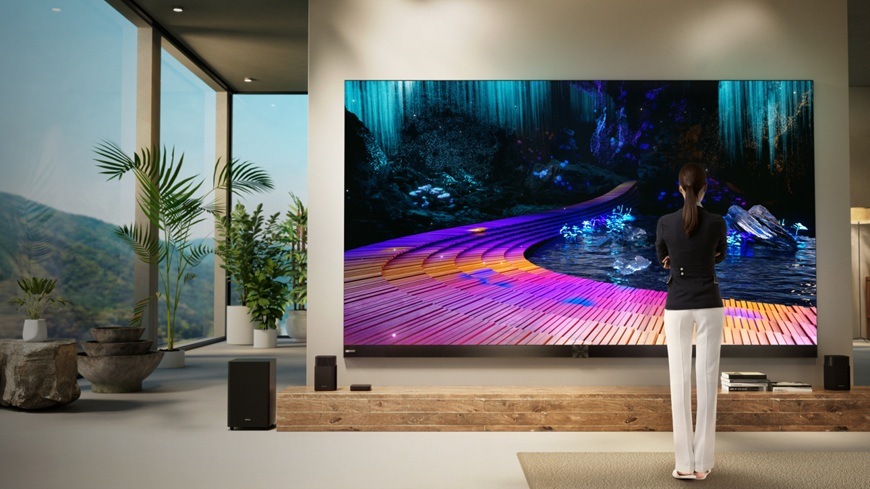 televize Hisense 136MX umístěná v moderním obývacím prostoru zobrazuje výrazně barevnou scénu s vysokým kontrastem a detaily. Postava stojící před obrazovkou podtrhuje impozantní velikost displeje.