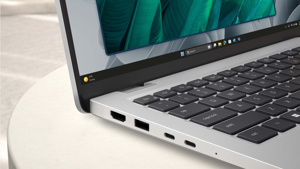 Elegantní notebook leží otevřený na světlém povrchu a na jeho zářivém displeji se zobrazují zelené listy. Stříbrné tělo je vybaveno porty USB a HDMI.