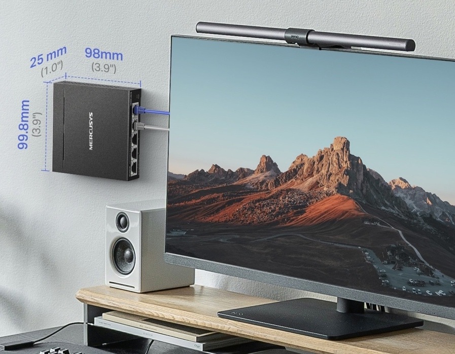 Switch Mercusys připevněný na stěně vedle monitoru. Obrázek uvádí kompaktní rozměry 99,8 × 98 × 25 mm.