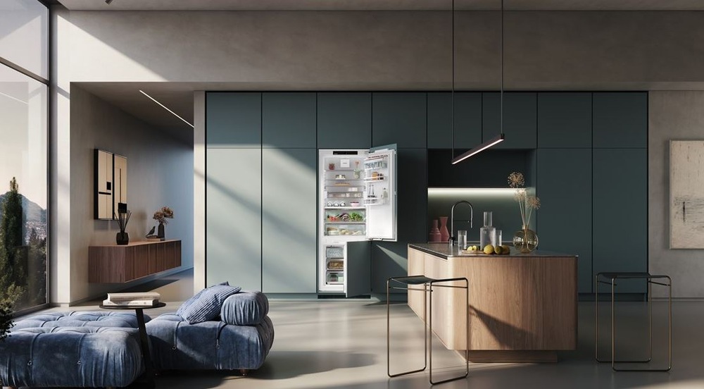 lednice Liebherr ICBNbi 7663 zabudovana v tmavě zelené kuchyni, zboku svíti slunce a vrha na skřínky stín. Lednice je otevřená a plná jidla.