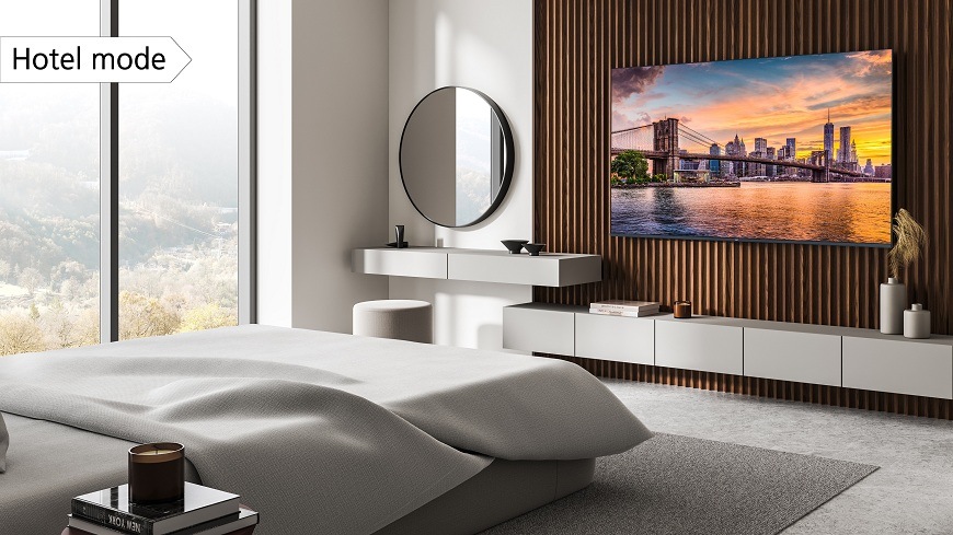 Televize JVC LT-65VDQ9435 JVC TV v moderním, světlém hotelovém pokoji a velkou postelí. Na televizi je zobrazen západ slunce nad Brooklynským mostem v NYC. V levém horním rohu je popisek 'Hotel mode' zdůrazňující režim pro hotely