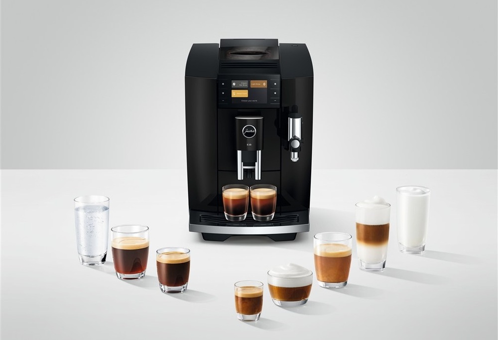 Automatický kávovar Jura E8 Piano Black připravuje dva šálky kávy zároveň, kolem kávovaru je rozestavěno osm sklenic s různými druhy kávy
