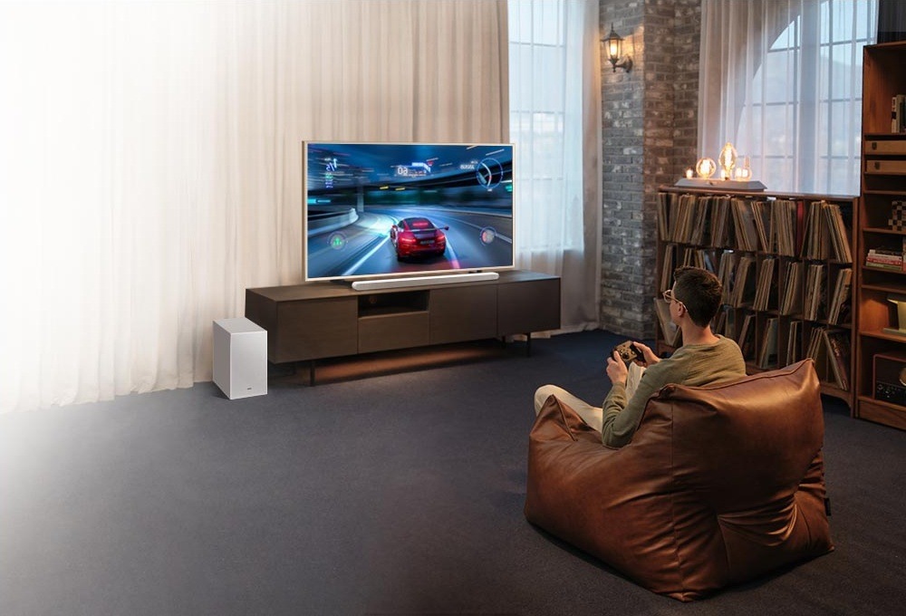 Soundbar Samsung HW-S701D bílý Muž sedí na sedacím vaku na zemi před televizí a soundbarem Samsung HW-S701D na televizním stolku a hraje videohru