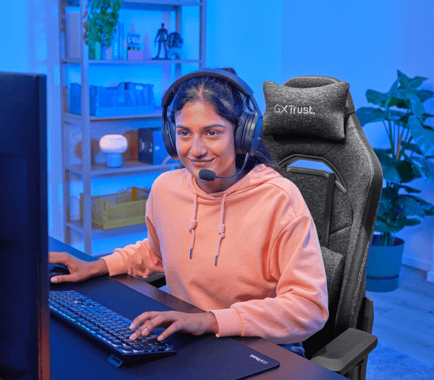 Žena se sluchátky sedící u herního stolu na židli Trust GXT 723 RUYA Fabric