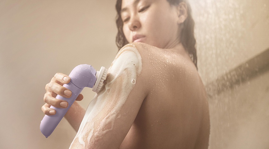 Žena používá epilátor Philips BRE728/00 ve fialové barvě s exfoliačním kartáčem na mokré pokožce ramene, které je pokryté pěnou během péče o tělo.