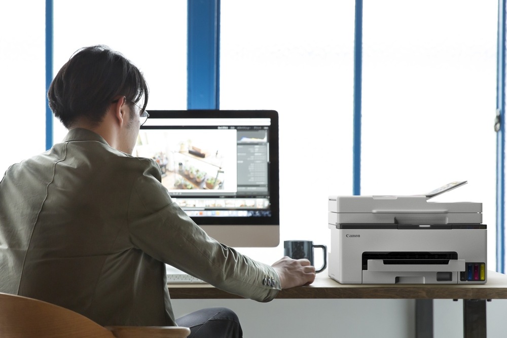 Tiskárna multifunkční Canon MAXIFY GX2041 bílá Muž pracující u monitoru s tiskárnou Canon MAXIFY GX2040 na stole v kanceláři.