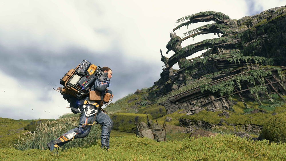 Hlavní hrdina hry Death Stranding s objemným batohem kráčí po travnatém svahu směrem k zřícené budově porostlé mechem pod zataženou oblohou.