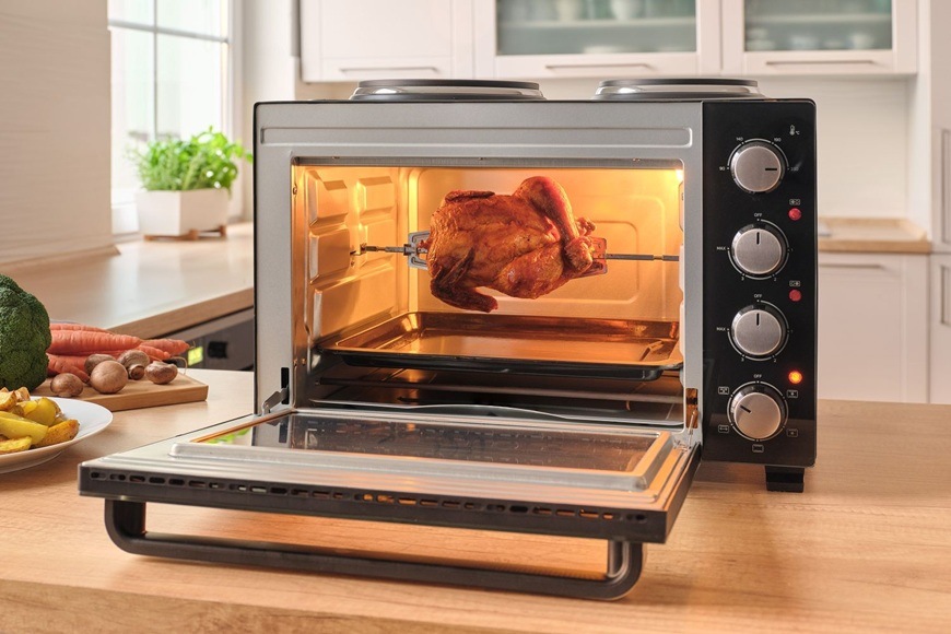 Otevřená mini trouba Sencor SEO 2800BK s otočným rožněm při pečení celého kuřete, v moderní kuchyni s nasvíceným vnitřkem a připravenou zeleninou na pracovní desce.