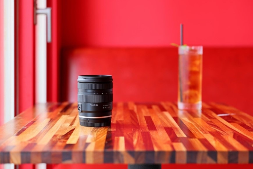 Objektiv Tamron 11-20 mm F/2.8 Di III-A RXD (Canon RF-S) černý Zátiší s nápojem a objektivem Tamron 11–20 mm F/2.8 Di III-A RXD na stolku v kavárně