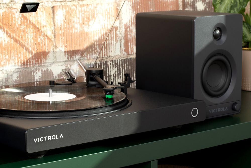 Detail gramofonu Victrola VPT-1520 Wave přehrávající vinyl, připojený k externímu reproduktoru na polici v domácím prostředí
