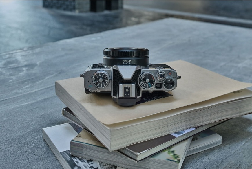Objektiv Nikon NIKKOR Z 26 mm f/2.8 černý Fotoaparát s objektivem NIKKOR Z 26 mm f/2.8 položený na hromádce knih