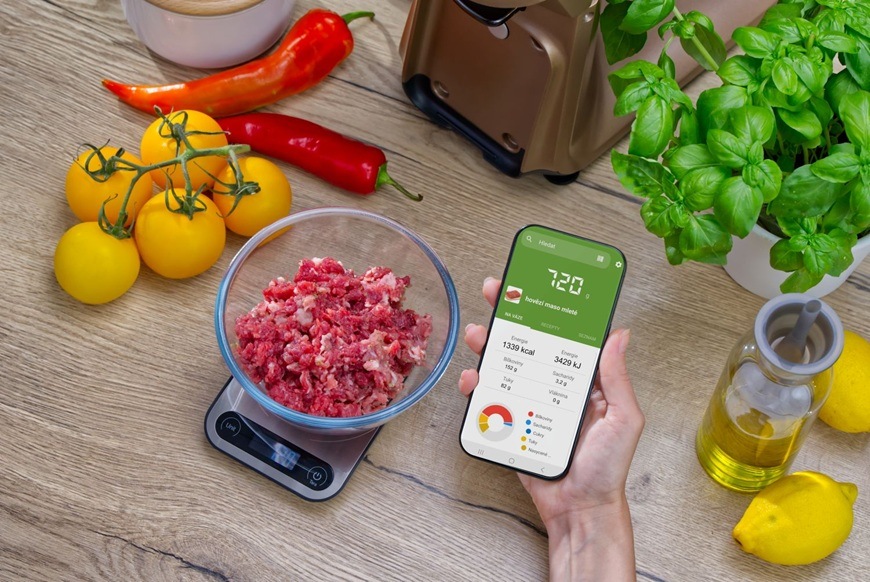 Žena drží v ruce telefon a v něm má otevřenou aplikaci Sencor FOOD, která ukazuje výživové hodnoty připravovaného pokrmu.