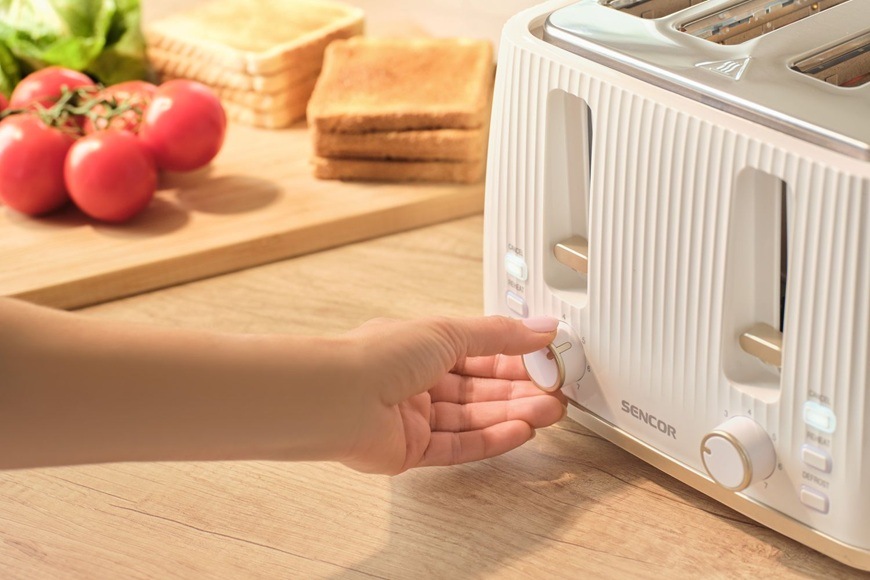Topinkovač Sencor STS 7540WH bílý Žena otáčí knoflíkem na stylovém topinkovači Sencor STS 7540WH v bílém provedení. V pozadí jsou na prkénku položené topinky a rajčata.