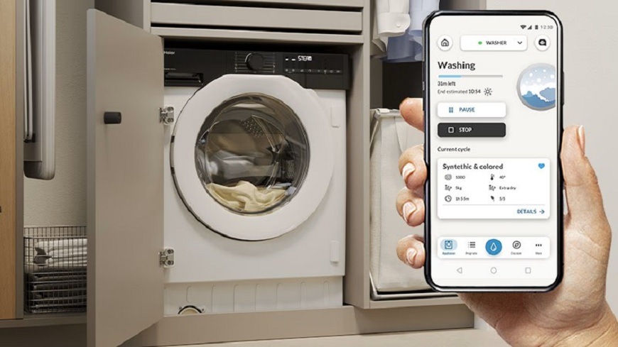 Pračka Haier BHA6S69M6DB9J-S bílá ruka drží telefon se zapnutou aplikací hon, v pozadí je pračka Haier BHA6S69M6DB9J-S s prádlem