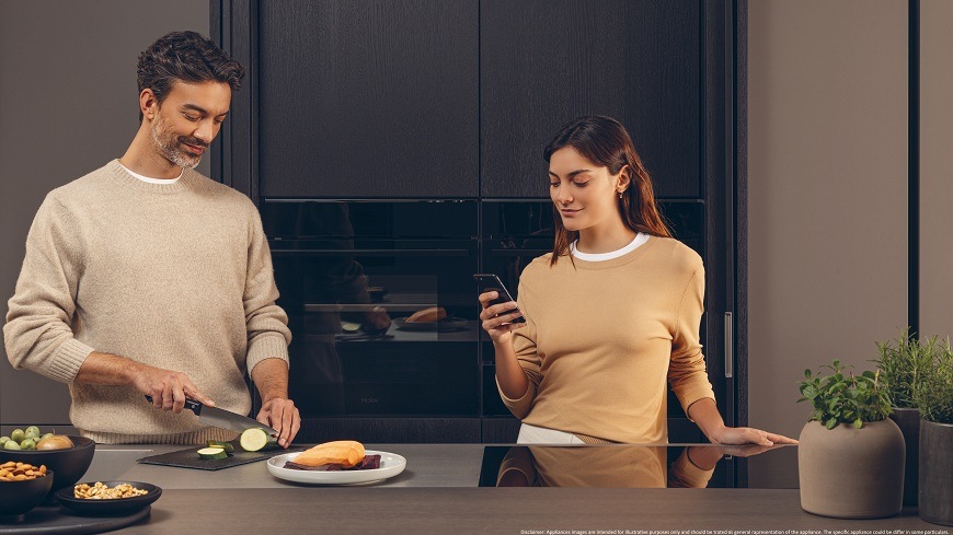 trouba Haier H6 ID2P3B4VYTB dva lidé jsou u kuchyňské linky, muž krájí cuketu, žena pomocí mobilu ovládá troubu, kterou mají za sebou